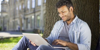 多种族大学生坐在树下，手提电脑，检查期末论文
