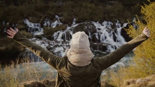 年轻美丽的女人站在瀑布附近，举起双手，感觉自由和幸福视频素材模板下载