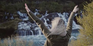 年轻的旅行女子站在冰岛瀑布附近微笑着。女孩举起手，感到高兴