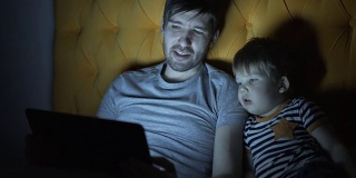 年轻的父亲和他的小儿子晚上躺在床上看卡通电影