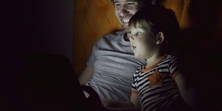 年轻的父亲和他的小儿子晚上躺在家里的床上用平板电脑看卡通电影和聊天