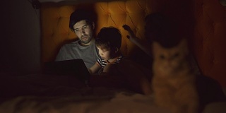 快乐的家庭与小儿子学习玩平板电脑躺在床上与猫在家里