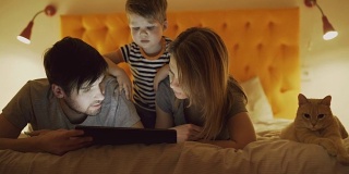 快乐的家庭和小儿子和有趣的猫躺在家里的床上，在睡觉前用平板电脑玩游戏