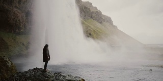 一名年轻男子带着摄像机站在冰岛的Gljufrabui瀑布附近，拍摄水流的视频