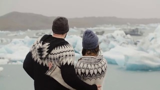 年轻夫妇站在冰泻湖的后视图。男人亲吻女人，拥抱她，看着冰岛的冰川视频素材模板下载