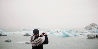 年轻的旅行男子站在冰岛的Jokulsalon冰礁湖，用智能手机拍摄冰川的背影