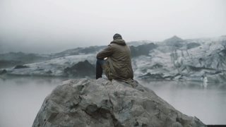 后视图的年轻游客坐在岩石上，看着冰川泻湖。独自探索冰岛的人视频素材模板下载
