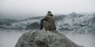 后视图的年轻游客坐在岩石上，看着冰川泻湖。独自探索冰岛的人