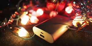 银色powerbank外部电池，用于在黑暗中放置在桌子上的智能手机或平板电脑，周围环绕着闪烁的花环灯