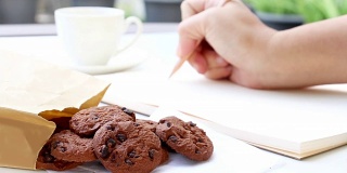 一名女子用一支木笔在笔记本上写着字，放在一张白色的桌子上随手拿着吃了一袋巧克力饼干和喝了一杯咖啡。