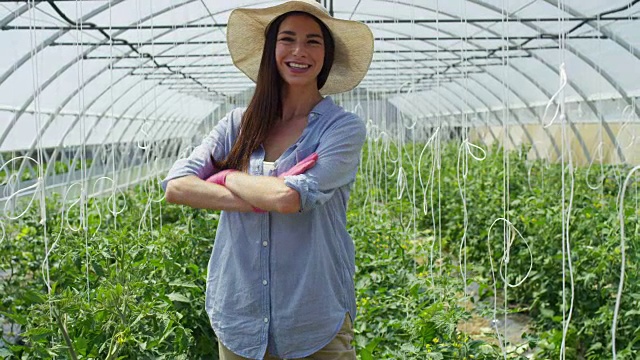 美丽的女孩戴着草帽，戴着粉色橡胶手套，微笑着，在一个温室里。概念:生物产品、天然产品、新鲜、美味、水果、蔬菜、种植、水、植物。