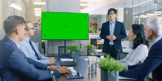 亚洲商人向他的商业伙伴做报告/报告，指着绿色度键屏幕墙电视。