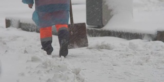 冬天。下雪的时候，一个工人穿着工作服拿着铲子在清理积雪。冬天在这个城市的街道上倒塌了。腿部特写