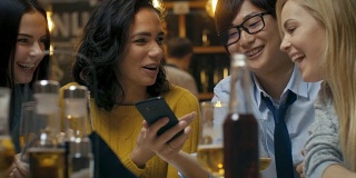 当朋友们在酒吧玩得开心时，年轻的亚洲男子在他的智能手机上向他们展示有趣的东西。他们在时尚的时髦酒吧里大笑，开玩笑，喝酒。
