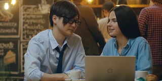 英俊的亚洲男人和美丽的高加索年轻女人坐在咖啡馆在笔记本电脑上工作。在后台其他客户在时尚的环境。