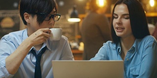 英俊的亚洲男人和美丽的高加索年轻女人坐在咖啡馆在笔记本电脑上工作。在后台其他客户在时尚的环境。