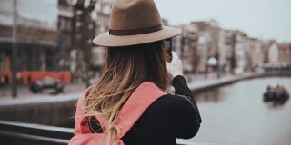 戴着帽子的年轻女孩在风景优美的河边拍照。一个留着长发背着红色背包的女孩在她的博客上分享照片。4 k