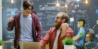 两位创意设计师看到笔记本电脑并取得了巨大成功，他们击掌并大笑。在有才华的人工作的背景时尚的办公室。