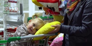 一位带着孩子的年轻妇女在超市选购商品。妈妈正用手推车载着一个小女孩。妈妈和女儿在看枕头。
