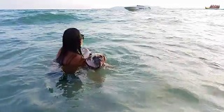 女人训练哈巴狗在海水里游泳，带着皮带和救生衣，有快艇和人在后面玩。