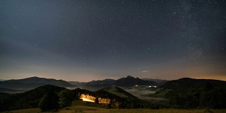 在月光下，星星在山峦和朦胧的乡村景色的夜空中移动。从黑夜到白天的时间流逝摄影车拍摄