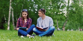 学生们正坐在草地上，一个男人在看书，一个女人在用平板电脑
