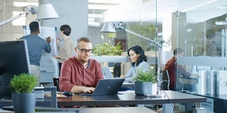 忙碌的国际公司办公室，白种人在他的办公桌上用笔记本电脑工作。在背景创造性的年轻人工作。