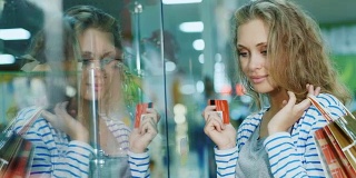 迷人的年轻女子在看一个玻璃展示柜在商店。拿着购物卡和购物袋的手