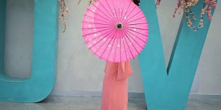 穿着粉红色和服的艺妓带着雨伞在墙边走着，上面写着樱花的大字母。