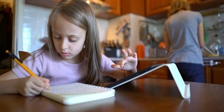金发女孩在笔记本上写字，在平板电脑上浏览。