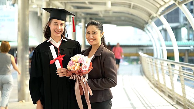 美丽的微笑毕业的学生女孩的肖像年轻的女人和她的母亲在帽子长袍走和拿着文凭卷轴。