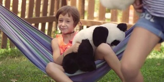年轻快乐的小女孩在夏令营躺在彩色的吊床上放松地玩在外面的毛绒狗娃娃在绿色的草地上孩子们，孩子们的慢动作
