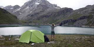 无人机拍摄，一名年轻女子在徒步旅行站在她的帐篷附近美丽的山湖双臂伸展