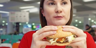 饥饿的女孩在美食广场吃汉堡。在快餐店里咬着芝士汉堡的女人