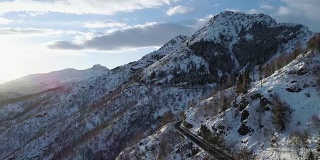 在靠近森林森林的冬季雪山上行驶的汽车的前空中俯视图。雪山街道道路的建立。日落或日出时太阳背光。全高清无人机飞行建立商店