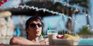 年轻英俊的男人戴着墨镜，喝着鸡尾酒，吃着热带水果，在旅游度假的游泳池里放松。1920 x1080