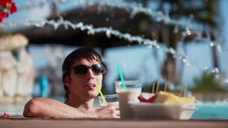 年轻英俊的男人戴着墨镜，喝着鸡尾酒，吃着热带水果，在旅游度假的游泳池里放松。1920 x1080视频素材模板下载
