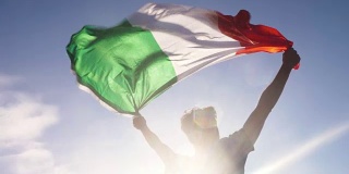 夕阳西下的意大利海滩上，一个年轻人双手举着意大利国旗向天空飞翔