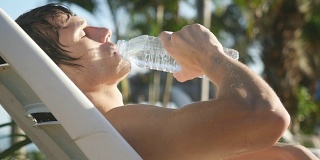 在泳池边晒太阳的帅哥从瓶子里喝水，慢动作。1920 x1080