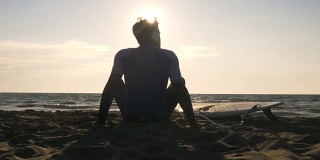 年轻的冲浪者穿着潜水服，坐在冲浪板上，躺在沙滩上，听着音乐，跳着舞，看着日落时的大海