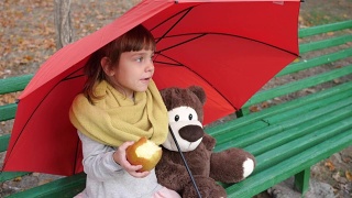 孤独的小女孩坐在伞下的木凳上，旁边是泰迪熊，在大自然的秋天吃着红苹果视频素材模板下载