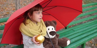 孤独的小女孩坐在伞下的木凳上，旁边是泰迪熊，在大自然的秋天吃着红苹果