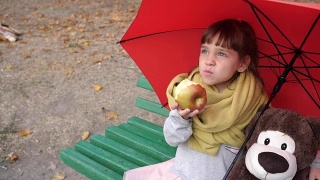 小女孩和玩具朋友坐在公园的一把伞下的长椅上吃红苹果视频素材模板下载
