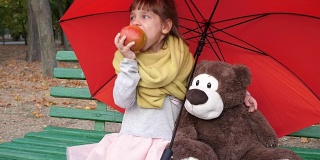 孩子们坐在红色雨伞下的木凳上，旁边是泰迪熊，在大自然的秋天吃苹果