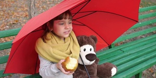 可爱的小女孩在大自然的伞下的木凳上吃着大的红苹果，旁边是泰迪熊