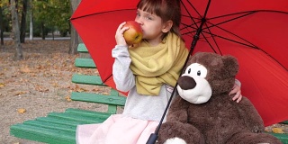 孩子坐在木凳上，旁边是一把红色伞下的泰迪熊，在公园里吃大红