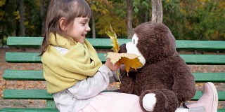 女孩坐在长椅上，手里拿着黄叶，背景是秋季公园里的泰迪熊