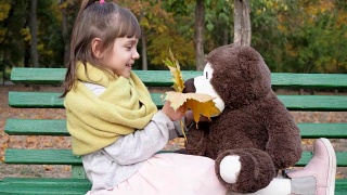 女孩坐在长椅上，手里拿着黄叶，背景是秋季公园里的泰迪熊视频素材模板下载