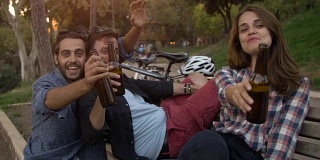 三个年轻的朋友，游客与自行车坐在长凳上的罗马斗牛场下的树在日落喝啤酒，有乐趣，聊天，大笑在罗马的慢动作