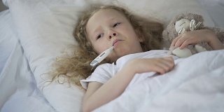生病的小女孩伤心地躺在床上，嘴里叼着温度计，孩子感冒了